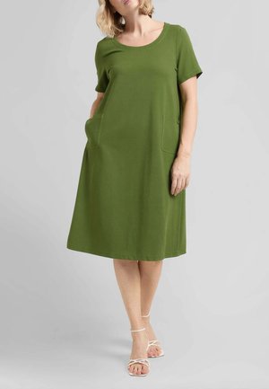 Donna che indossa un vestito verde oliva a maniche corte, lunghezza ginocchio, con due tasche frontali e sandali bianchi con cinturini, in piedi davanti a uno sfondo semplice.