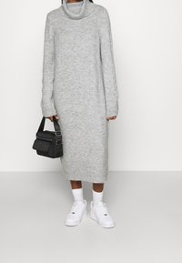 Abito maglione grigio con collo a lupetto, maniche lunghe e orlo a coste. Abbinato a sneaker bianche e una piccola borsa a tracolla nera.