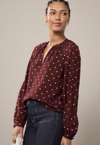 Blusa burdeos con lunares blancos, de mangas largas con puños elásticos y un escote en V profundo. Textura suave, tejido ligero, corte casual.