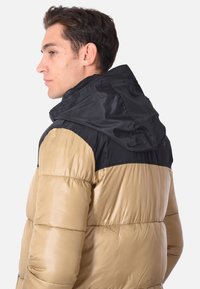 Giacca piumino con design bicolore: cappuccio e parte superiore neri, con corpo inferiore color sabbia. Texture liscia e lucida, con polsini elasticizzati.