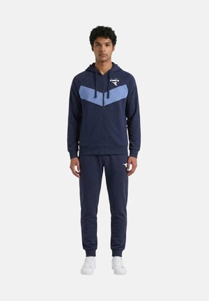 Uomo con tuta blu scuro con motivo a chevron azzurro chiaro sulla giacca e scarpe da ginnastica bianche, in piedi di fronte a sfondo bianco.