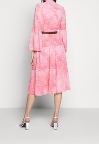 Robe rose à motif cachemire, manches longues évasées, taille cintrée et jupe fluide. Associée à des sandales blanches à talons blocs.