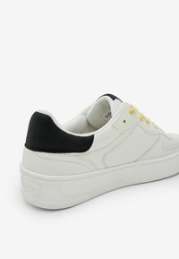 Sneaker bianco con tallone in suede nero, lacci gialli, tomaia in pelle liscia e suola in gomma testurizzata.