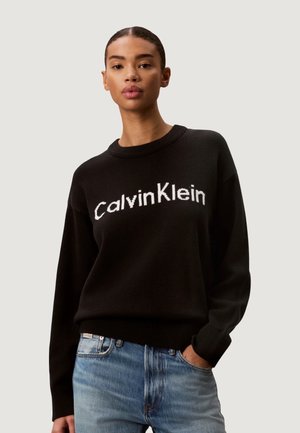Kobieta nosząca czarny sweter Calvin Klein i niebieskie dżinsy, stojąca z jedną ręką w kieszeni na jednolitym tle.