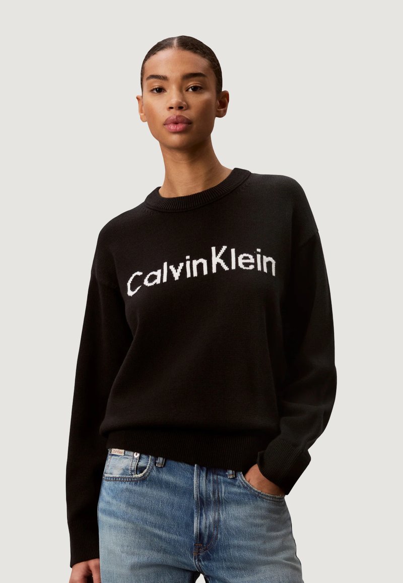 Femme portant un pull noir Calvin Klein et un jean bleu, debout avec une main dans la poche devant un fond uni.