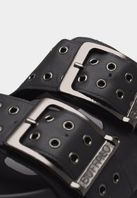 Κοντινό πλάνο από λουριά μαύρων δερμάτινων σανδαλιών με ασημένιες μεταλλικές αγκράφες και στρογγυλές τρύπες, μία αγκράφα χαραγμένη με το όνομα της μάρκας «BUFFALO».