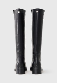 Bottes en cuir noir arrivant au genou avec une texture lisse, bout arrondi et petit talon carré. Présente de petites accents argentés en haut.