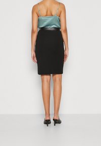 Svart pennkjol med en figurnära design, kombinerad med en ljus teal satin camisole och svarta klackar. Slät textur; minimalistisk stil.