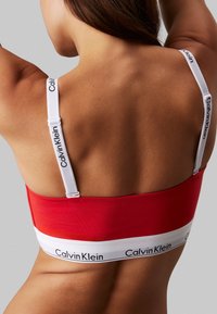 Rdeča bralette z belimi trakovi in blagovno znamko. Ima gladko teksturo, prilegajoč dizajn in podporni pas z detajlom logotipa.
