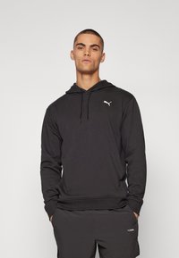 Ung mand iført sort Puma hoodie og matchende shorts, stående med hænderne i lommerne mod en ensfarvet baggrund.