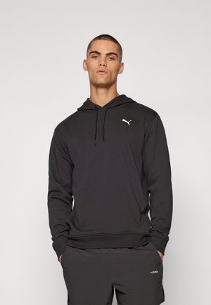 Ung mann iført svart Puma hettegenser og matchende shorts, stående med hendene i lommene mot en ensfarget bakgrunn.