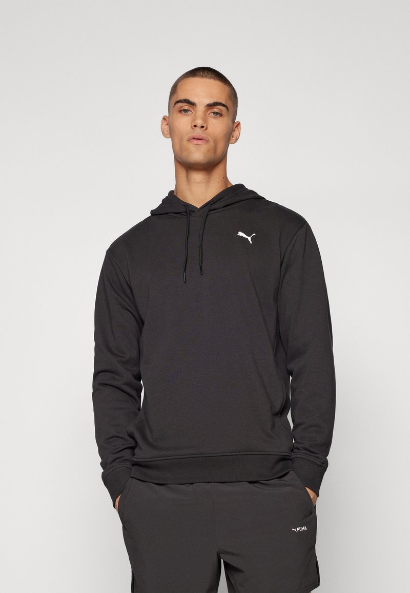 Ung mand iført sort Puma hoodie og matchende shorts, stående med hænderne i lommerne mod en ensfarvet baggrund.