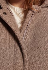 Cappotto marrone texturizzato con una cerniera frontale, dotato di un bottone a scatto e un maglione intrecciato color crema sottostante. Primo piano dei dettagli del tessuto.