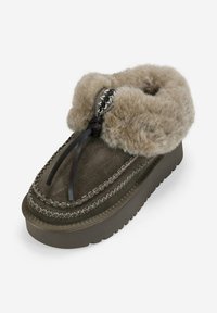Mocasines de ante marrón con suela gruesa de plataforma, cuello de piel beige suave, costuras decorativas y lazo de cuero en un fondo blanco.