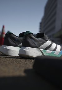Coppia di scarpe da ginnastica bianche e nere con dettagli verdi posate sull'asfalto vicino a coni stradali e un edificio urbano.