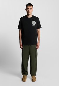 T-shirt en coton noir avec un graphique circulaire et du texte blanc, associé à un pantalon cargo vert olive ample et des chaussures en cuir suédé marron.