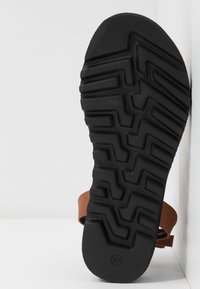 Ruskea sandaali paksulla mustalla kumipohjalla, jossa on viivastomalli. Pohja on yksityiskohtainen ja suunniteltu tarjoamaan pitoa. Koko merkittynä 37.