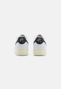 Sapatilhas Nike Air brancas com um detalhe azul marinho no calcanhar, logotipo dourado e sola com manchas em creme, exibindo um design clássico de cano baixo.