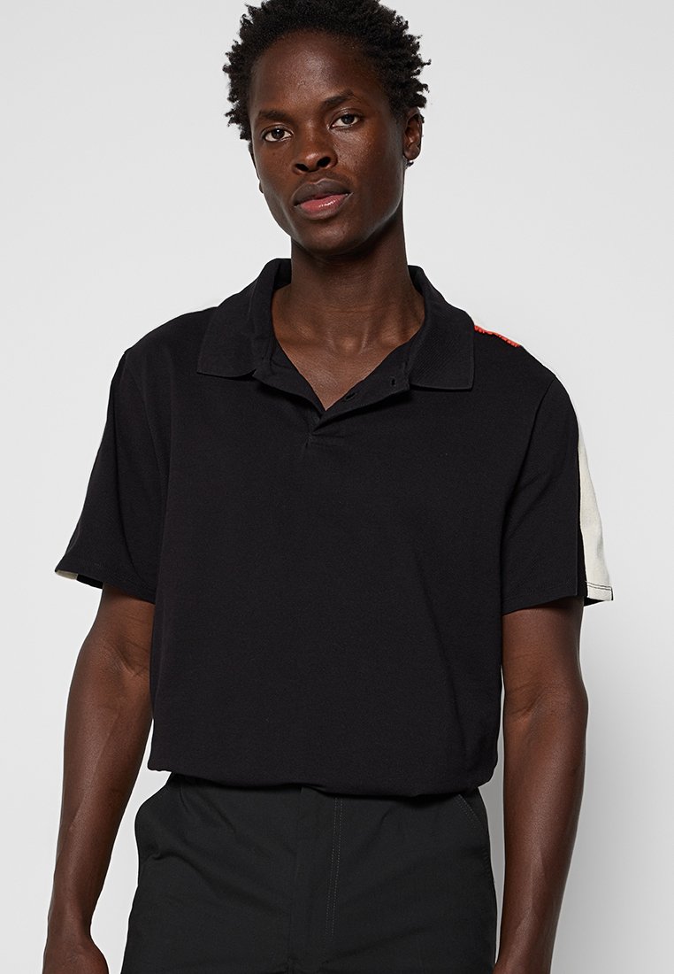 Polo noir avec un col, doté de manches courtes avec des accents blancs et rouges sur les épaules. Fabriqué en tissu de coton texturé.