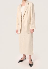 Blazer beige con rever a lancia, chiusura a un bottone e due tasche frontali, abbinato a una gonna su misura coordinata e scarpe con tacco nere.