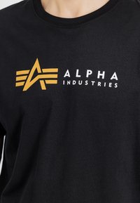 Μαύρη βαμβακερή μπλούζα με χαρακτηριστικό κίτρινo λογότυπο που διαθέτει τρεις οριζόντιες ρίγες και το κείμενο "ALPHA INDUSTRIES" σε λευκό χρώμα.
