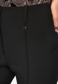 Schwarze, maßgeschneiderte Hose mit glatter Textur, schlanker Passform, Seitentaschen und einem kleinen schwarzen Knopfverschluss. Minimalistisches Design mit klaren Linien.