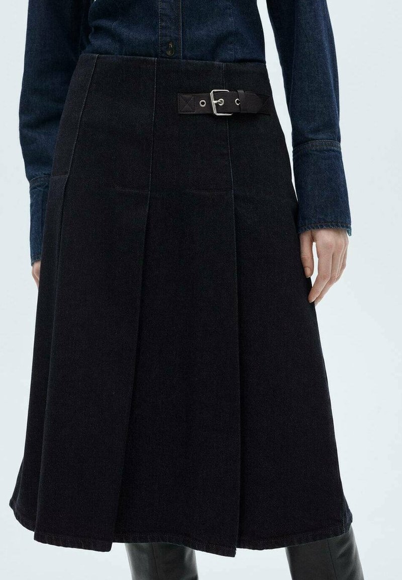 Jupe trapèze sombre avec boucle latérale, portée avec une chemise en denim foncé et des bottes noires montant jusqu'au genou, présentée en longueur moyenne sur une personne debout.