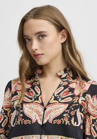 Blouse noire avec un imprimé cachemire et floral aux couleurs rouge, orange et blanc. Les détails notables incluent un devant boutonné et un col pointu.