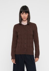 Brun kabelstrikket cardigan med ribbestrikket nederkant og ermer. Har svarte knapper og en liten logo på brystet. Båret over en stripete skjorte.