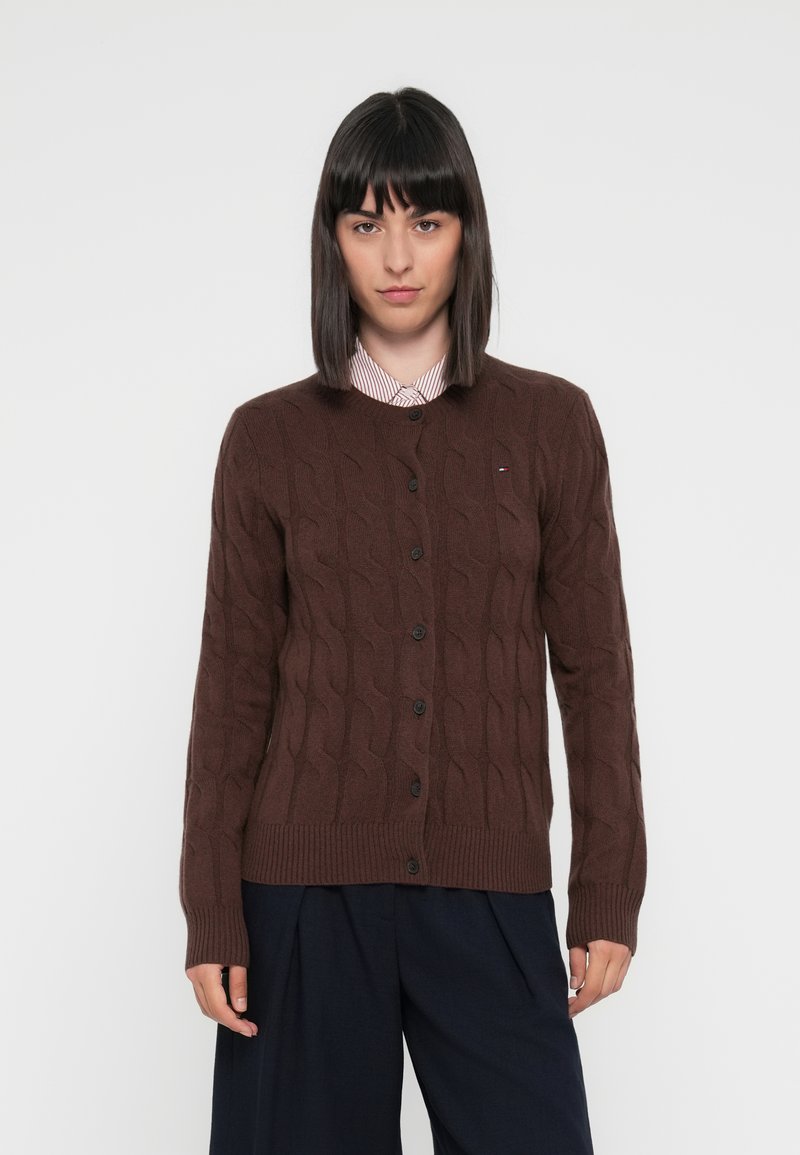 Brun kabelstrikket cardigan med ribbestrikket nederkant og ermer. Har svarte knapper og en liten logo på brystet. Båret over en stripete skjorte.