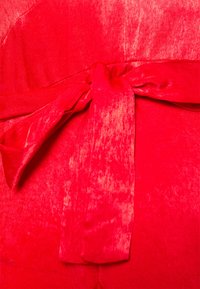 Vestido vermelho com uma superfície texturizada, apresentando um laço na cintura. O tecido tem um brilho ligeiro e um caimento suave, realçando a sua forma.