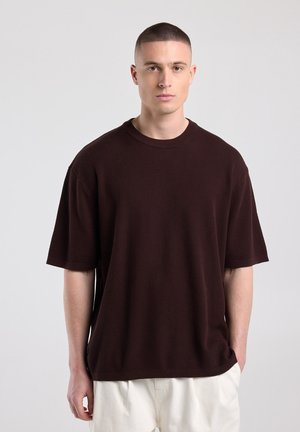 Jonge man met kort haar die een los donkerbruin shirt met korte mouwen en lichtgekleurde broek draagt, staand tegen een effen achtergrond.