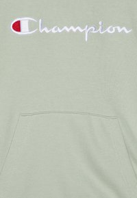 Felpa verde chiaro con tasca frontale, decorata con il logo "Champion" in corsivo bianco e un emblema rosso e bianco sulla sinistra. Texture morbida.
