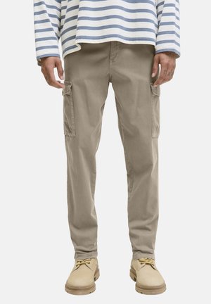 Hombre con pantalones cargo beige, botas marrones con cordones y una camisa blanca de manga larga con rayas horizontales azules.