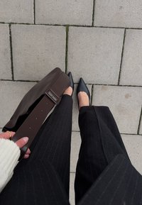 Person trägt schwarze Nadelstreifenhose und schwarze spitze Schuhe, hält eine große braune Bauchtasche über einem gepflasterten Steinweg.