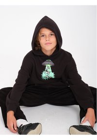 Zwarte hoodie met een grafisch ontwerp van een UFO en een lichtstraal, gecombineerd met zwarte corduroy broek en zwarte sneakers.