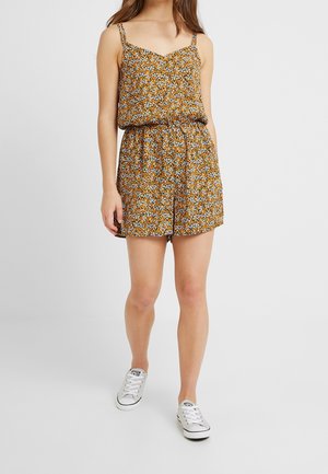 Femme portant une combinaison orange sans manches à motif floral avec des baskets blanches, debout devant un fond clair uni.