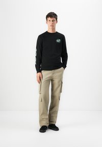 Jonge man die staat met één hand in de beige cargobroekzak, gekleed in een zwart lange mouwen shirt met turquoise tekst en zwarte schoenen.
