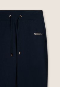 Joggers blu navy con vita a coste dotata di cordini, dettagli dorati e un logo discreto in lettere metalliche sulla coscia sinistra.