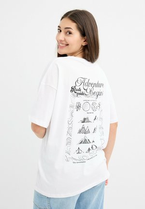 Mujer sonriente con cabello castaño liso que lleva una camiseta blanca oversized con ilustraciones de aventuras al aire libre en negro y texto en la parte de atrás.