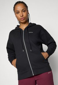 Čierna mikina na zips s podšívkou kapucne, prednými vreckami a logom DKNY. Má jemnú textúru a zips a akcenty v striebornom odtieni.
