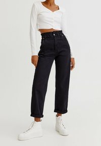 Vrouw in hoge zwarte paperbagbroek, witte cropped longsleeve top en witte platform veterschoenen, staand tegen een effen achtergrond.