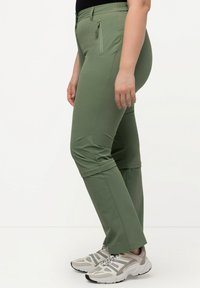 Pantaloni sportivi verdi con un design affusolato, vita elastica, tasca laterale con zip e orli arrotolati, abbinati a sneaker grigie.