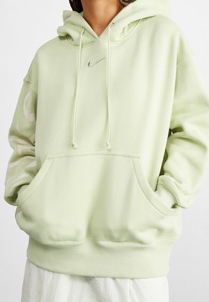 Osoba nosząca jasnozieloną bluzę z kapturem Nike z przednią kieszenią i troczkami, ręce wsunięte w kieszeń, widoczne białe spodnie poniżej.