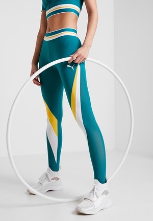 Vihreät urheiluhousut, joissa on keltaisia ja valkoisia viistoja raitoja, ja siihen sopiva lyhythihainen toppi, yhdistettynä valkoisiin tennareihin ja korvakoruihin.