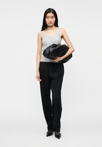 Sort læderhåndtaske med rundet form, båret af en person iført en grå ribbet tanktop og sorte bukser, mod en ensfarvet baggrund.