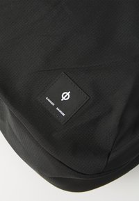 Gros plan sur un tissu noir avec un motif de grille texturé et une étiquette carrée noire comportant un cercle blanc avec une ligne diagonale et le texte "SAMSOE SAMSOE".