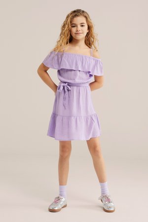 Jong meisje staand met handen achter de rug, gekleed in een lavendel kleurige off-shoulder jurk, bijpassende sokken en zilveren schoenen tegen een neutrale achtergrond.