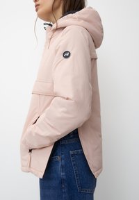 Hellrosa isolierte Jacke mit Kapuze, seitlichen Taschen, einem schwarzen Logo am Ärmel und einem etwas längeren Rückensaum.