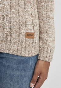 Beżowy sweter z dzianiny z wzorami warkoczowymi, ściągaczami na mankietach oraz skórzaną naszywką z logo „OXMO”. Noszony z niebieskimi dżinsami.